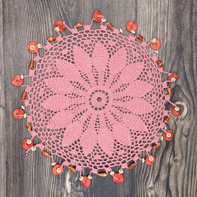 A round pink crocheted doily with a floral mandala pattern, decorated with small orange and white beads along the edge, laid flat on a dark gray wood plank surface.