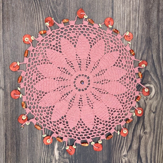 A round pink crocheted doily with a floral mandala pattern, decorated with small orange and white beads along the edge, laid flat on a dark gray wood plank surface.