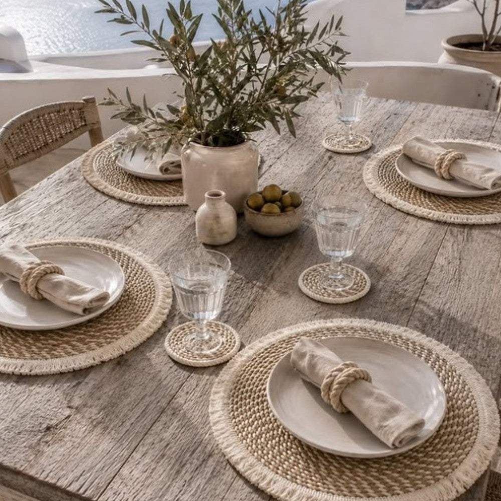 A styled dining table set with woven round placemats, white plates, beige napkins tied with rope rings, clear drinking glasses, a small ceramic vase with olive branches, and a bowl of olives in a bright outdoor setting.